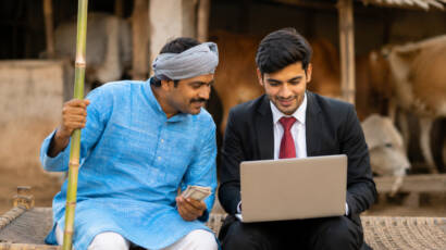 Rural banker discussing digital financial services with a farmer using a laptop in a village setting.